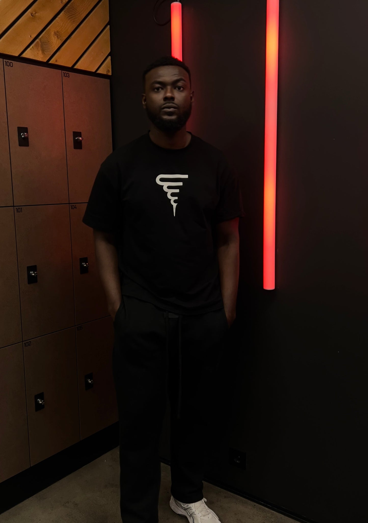 Person wearing a black t-shirt with a logo in a room with lockers and neon lights.